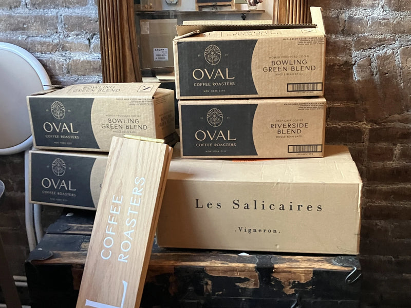 Oval Coffee Roasters wholesale master cases labeled Bowling Green and Riverside whole-bean coffee stacked against a brick wall beside a wooden Coffee Roasters sign and a wine box.